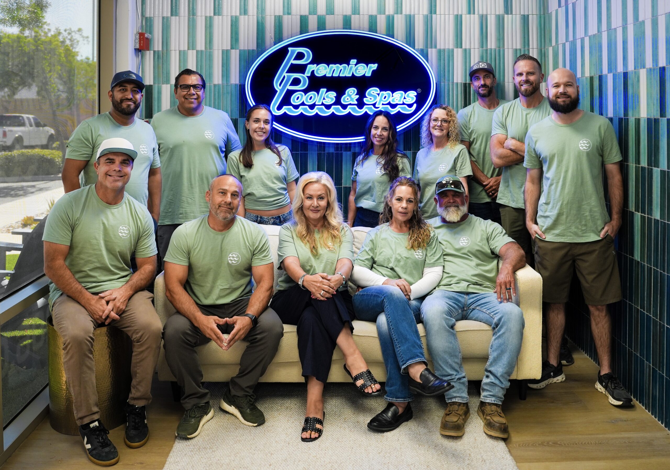 "Picture of the entire Temecula staff. 4 of them are sitting on a couch with a wall behind them displaying the Premier Pools and Spas logo. There are 9 other people standing around it."