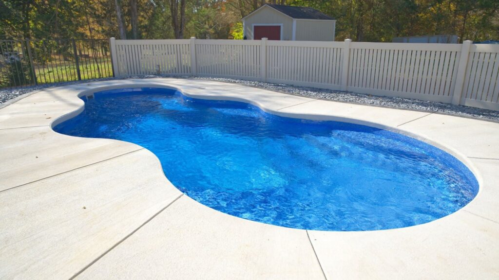 " Kidney-shaped inground pool with blue water, light concrete deck, and white vinyl fence"