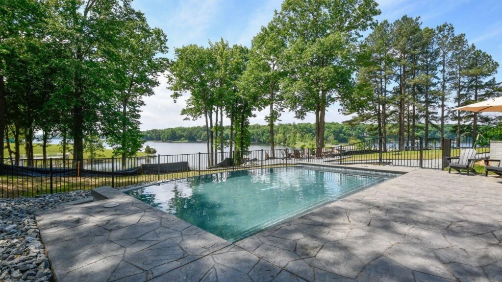 "Inground pool with stamped concrete deck and black fence, tall trees and a lake in the background"