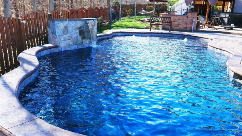 "Freeform inground pool with deep blue water, stone waterfall feature, and a wooden privacy fence"
