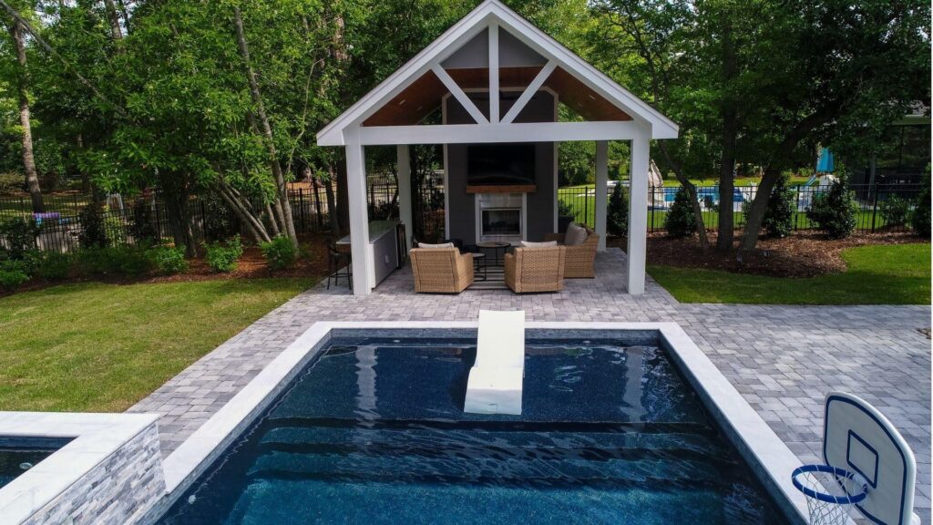 "Dark-bottom pool with a white tanning ledge, paver patio, and a pavilion with fireplace and wicker chairs"