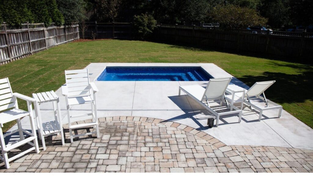 "Small rectangular blue pool with a concrete deck, white lounge chairs, and a paver patio in a fenced yard"