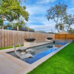 Narrow rectangular pool with sun-shelf lounge chairs, stone fire bowls, paver deck, and turf lawn