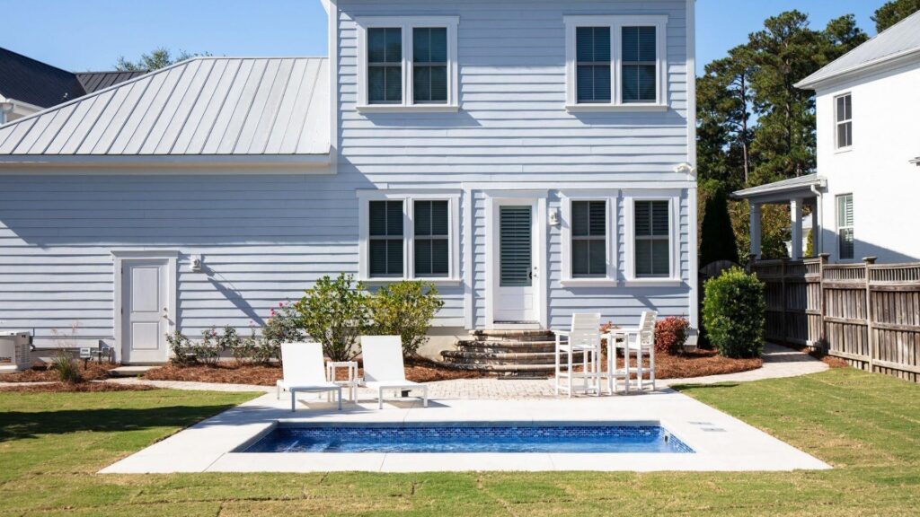 "Narrow rectangular plunge pool with white lounge chairs and bistro table behind a blue-gray house"