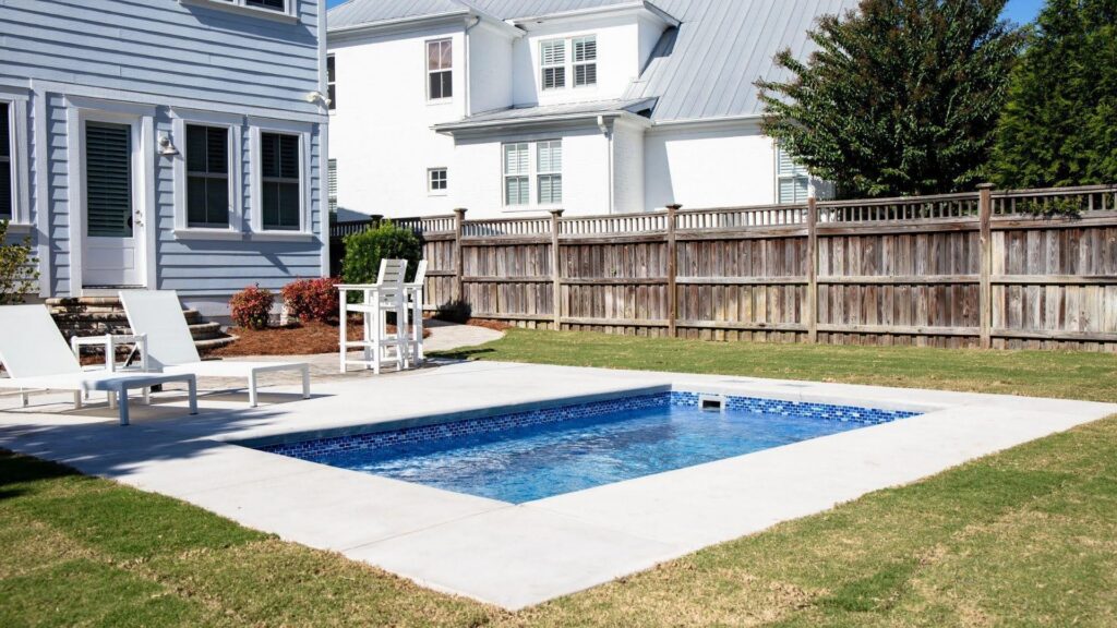 "Small rectangular pool with mosaic tile border, light concrete deck, lounge chairs, and wood fence"