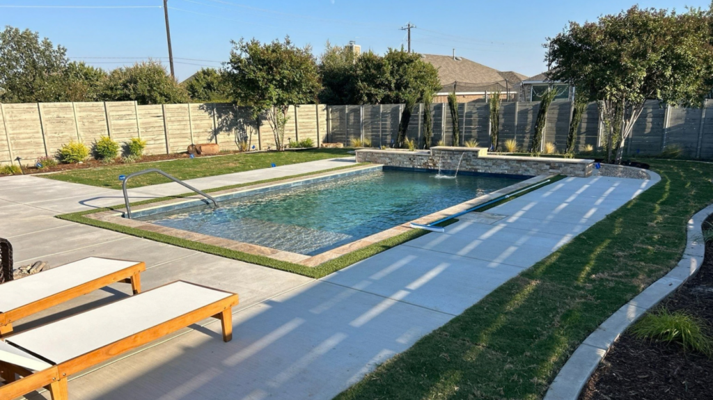 "Alt txt: Rectangular pool with raised stone water feature, tanning ledge, curved lawn path, wood fence"