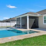 "Rectangular pool in a Jacksonville backyard with a sun shelf and entry steps, travertine deck and coping, a raised tiled wall with planters, and a screened sun room beside a white vinyl fence"