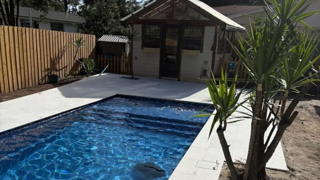 Rectangular inground pool with a light paver deck, wood privacy fence, and a small backyard cabana